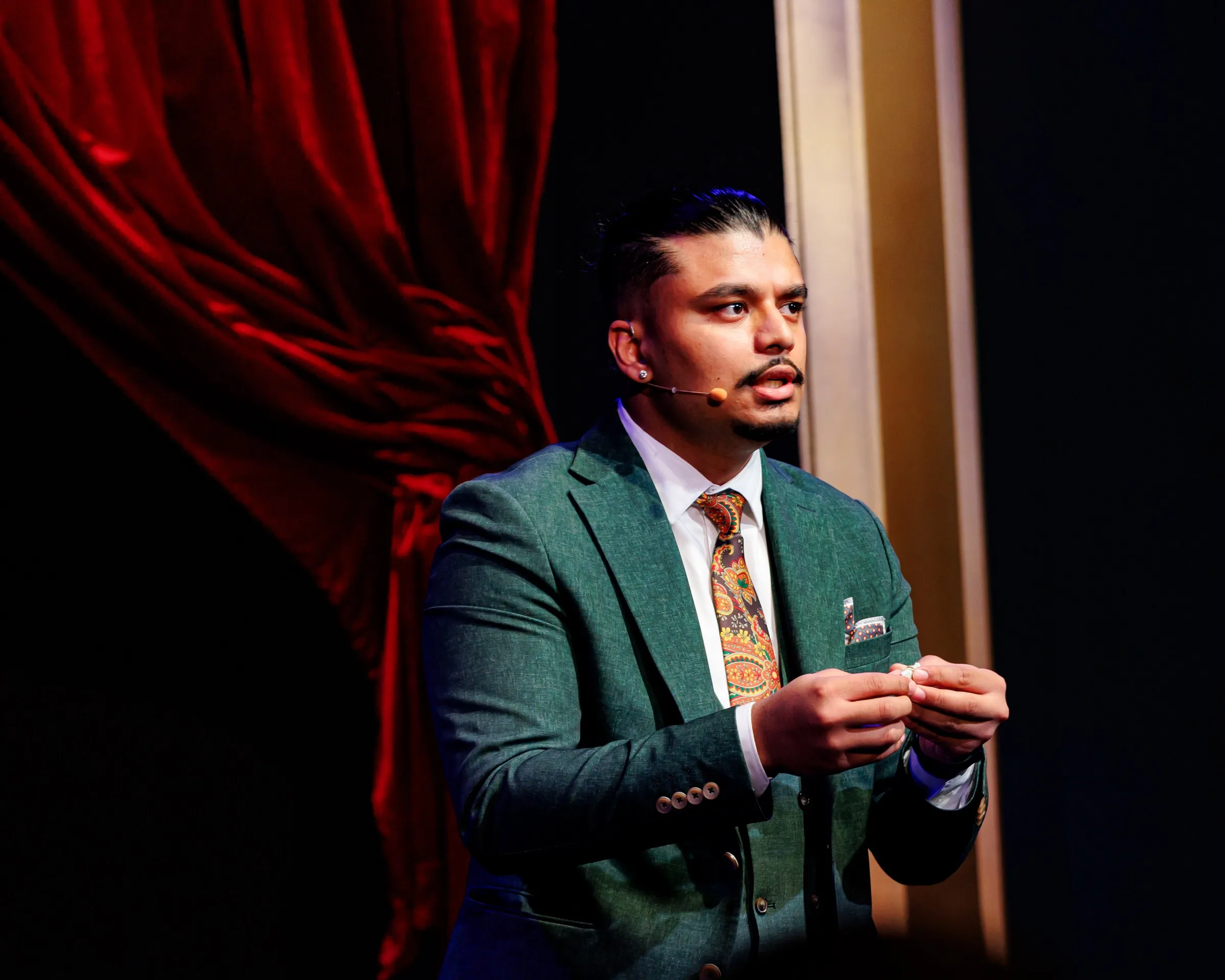 Savio Joseph on stage before a red velvet curtain.